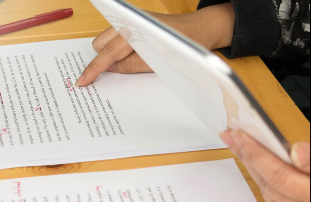 Image of a typed and annotated essay on a table; a student's hands holding a tablet and checking the paper are in the frame.