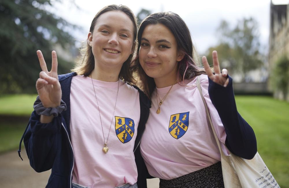 Two Trinity College female students wearing pink freshers committee t-shirts stand in the college's front quad making peace signs and smiling.