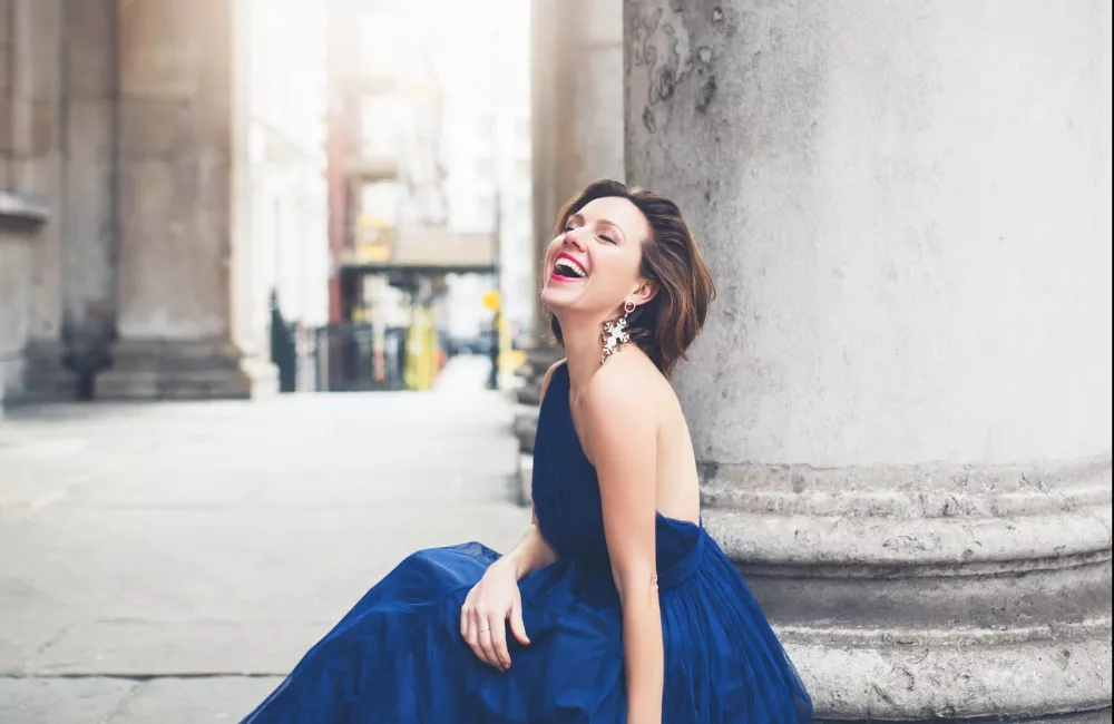 A woman in a blue ball gown sits on a the steps of a concert hall leaning against a column. 