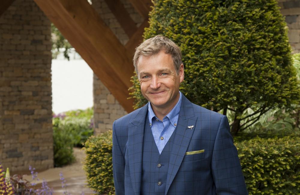 Chris Beardshaw stands smiling in a garden he designed; behind him is a large manicured tree. 