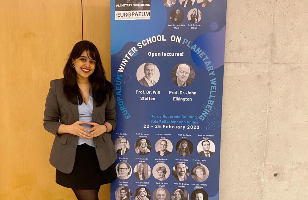Rai Sengupta stands in front of a banner advertising the Europaeum Winter School on Planetary Wellbeing.