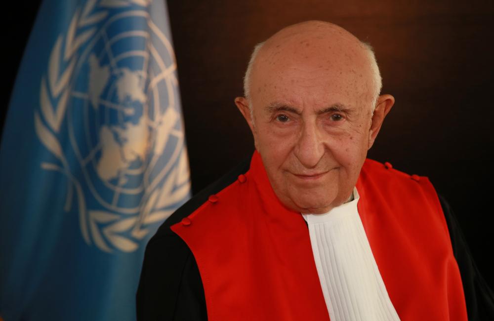 Judge Theodor Meron stands in his robes smiling in front of the flag of the United Nations.