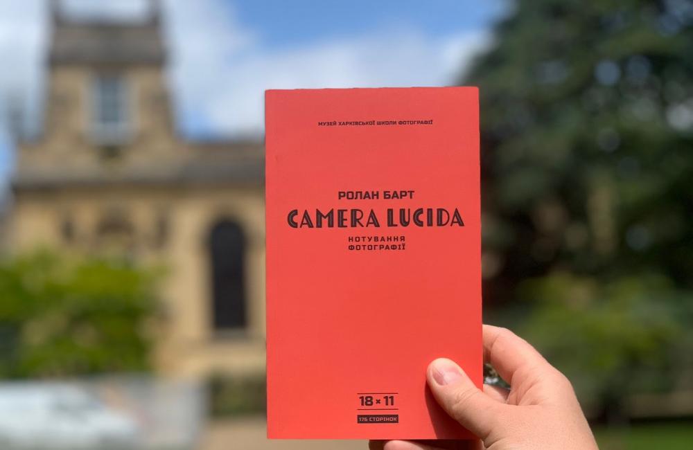 A hand holds up a copy of Camera Lucida in Ukrainian in front of the Trinity College chapel.