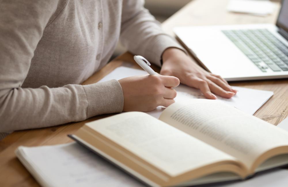 Shutterstock image of a person writing notes in a notebook with an open book and laptop next to them.