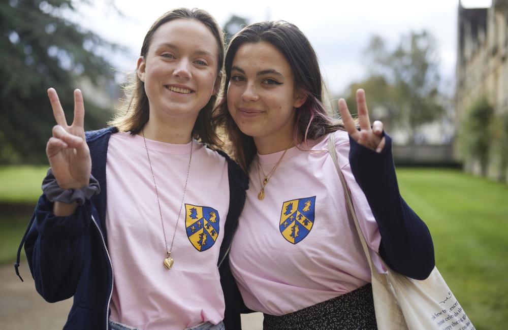 Two female Trinity students stand in the front quad of college making peace signs and smiling.