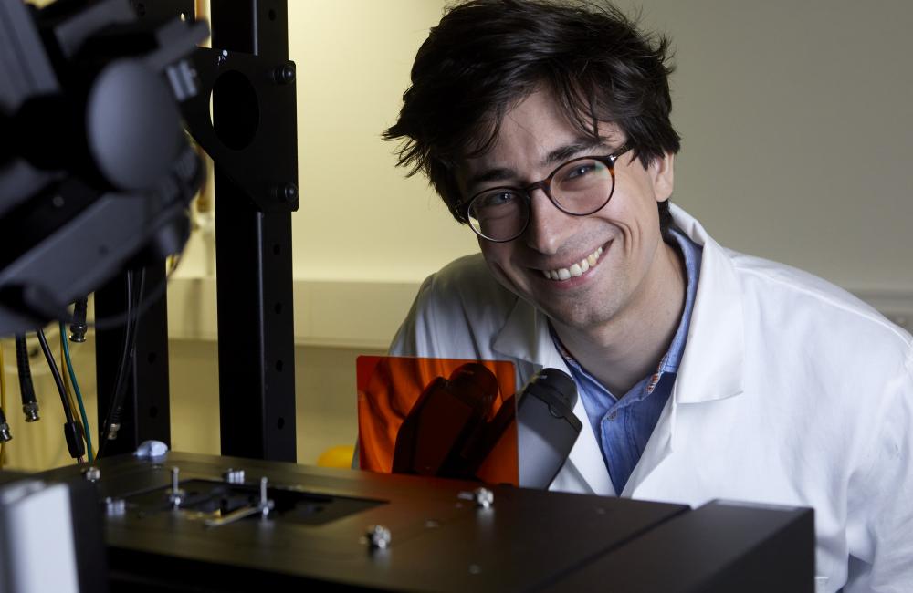 Trinity student Philippe Holzhey sits in front of a microscope; he is wearing a lab coat and goggles.