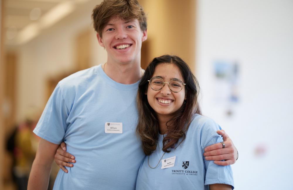 A male and female Trinity college student stand in matching student ambassador t-shirts with their arms around each other smiling.