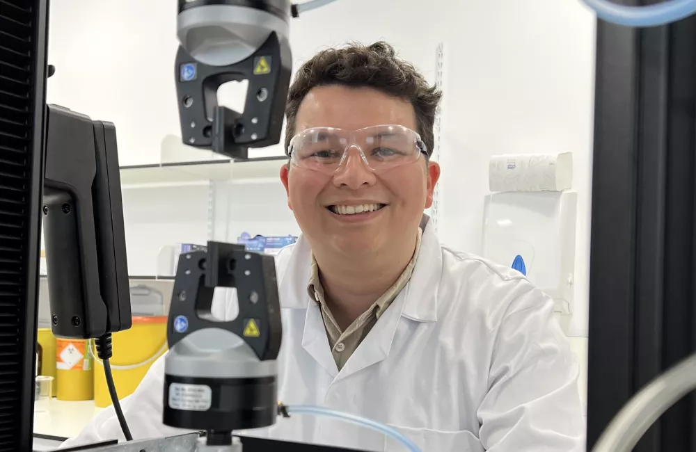 Trinity Dphil student Kam Poon sits at a lab microscope; he is wearing a white lab coat and goggles.