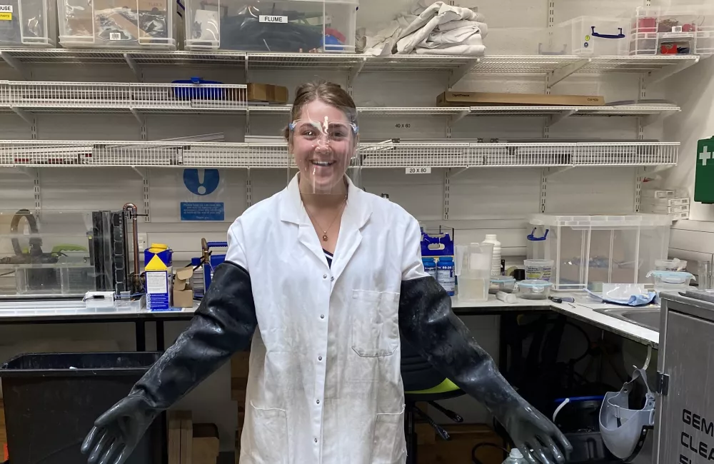 Trinity student Codrutza Dragu stands in the robotics lab wearing oversized lab coat and gloves, as well as safety goggles.