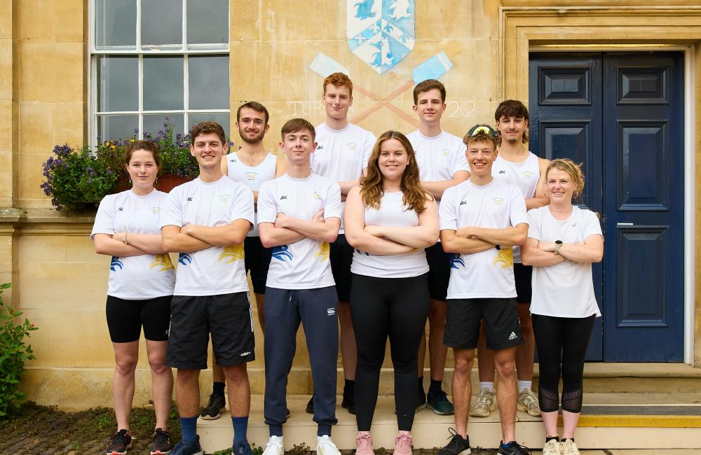Nine members of the Trinity College Boat Club stand in matching kit in front of a building with rowing oars chalked on.