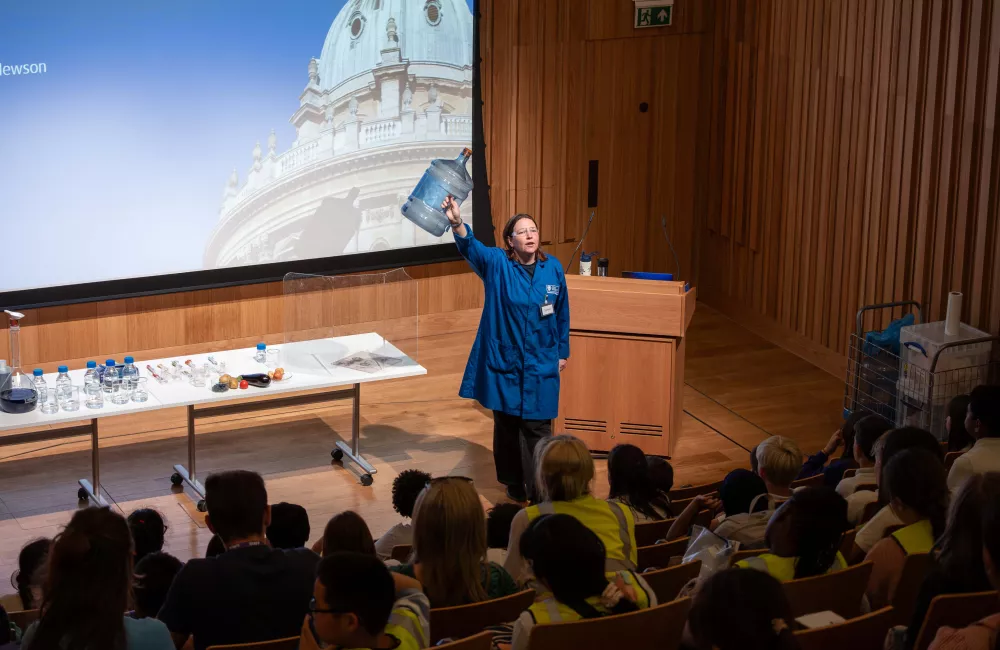 Science presenter in a blue lab coat holding up a large water container during a live demonstration in Trinity College’s de Jager Auditorium. A table with experiment materials is set up beside her, and a projected image of Oxford’s Radcliffe Camera is displayed behind. Young students in high-vis jackets watch attentively from tiered seating.
