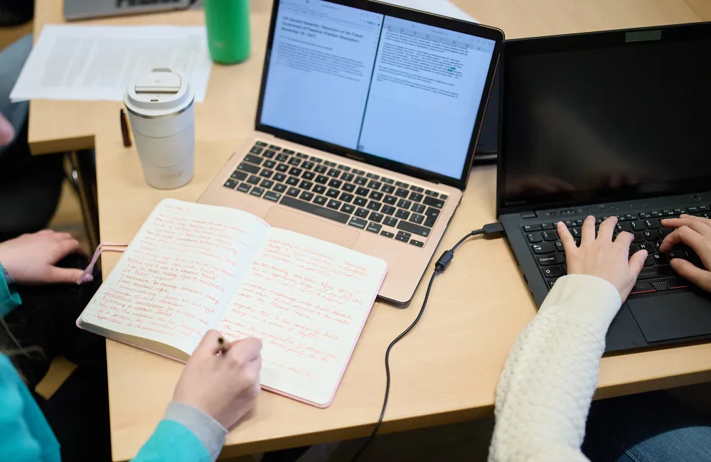 wo students working at a desk with laptops and an open notebook. One types on a black keyboard while the other takes handwritten notes in red ink. A coffee cup, water bottle, and printed document are also visible on the desk.