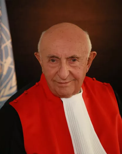 Judge Theodor Meron stands in his robes smiling in front of the flag of the United Nations.
