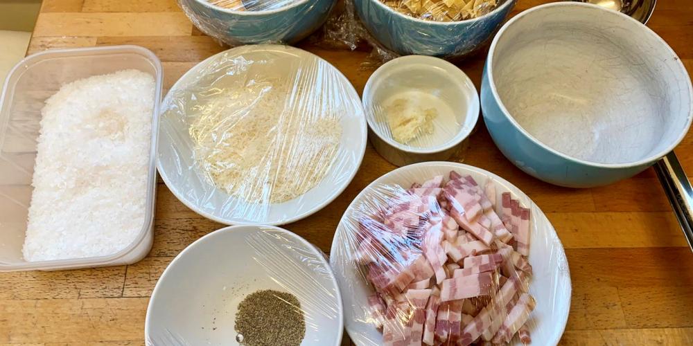 A table with bowls of pasta ingredients including bacon and cheese.