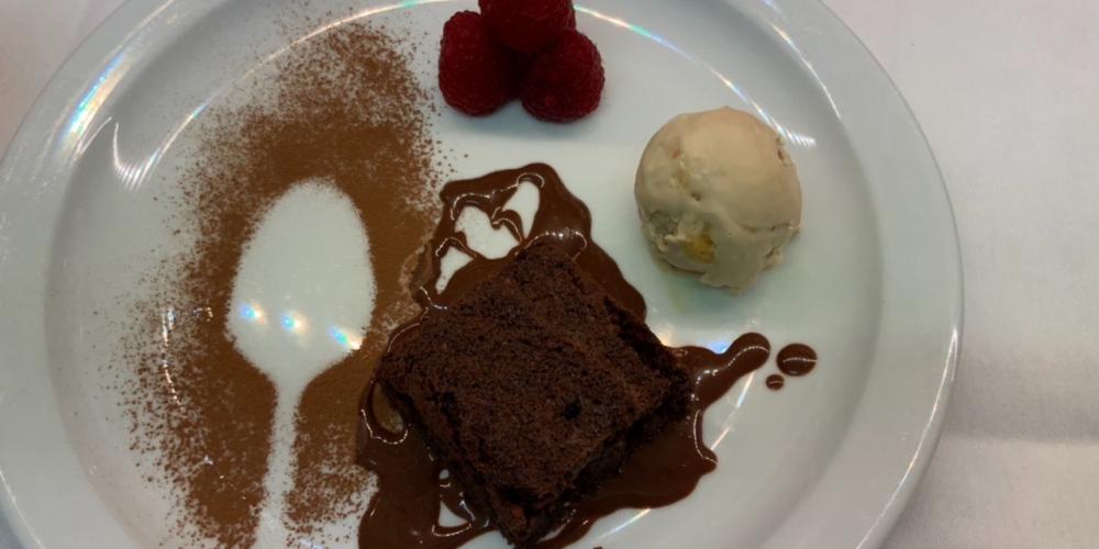 A plate with brownie drizzled with chocolate sauce and raspberries with a scoop of ice cream and sprinkled cocoa.