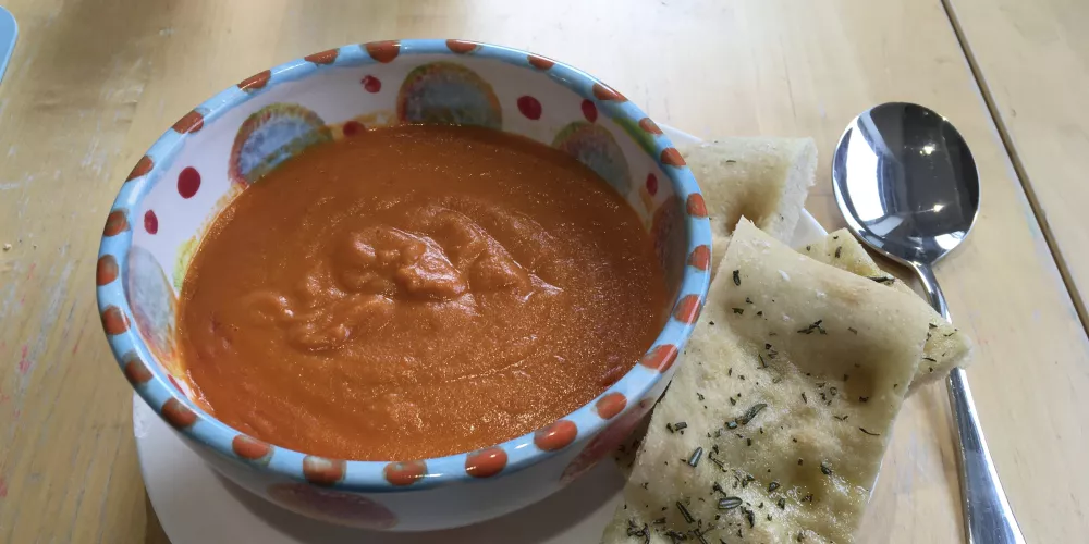 A bowl of tomato soup on a plate with a slice of focaccia bread. 