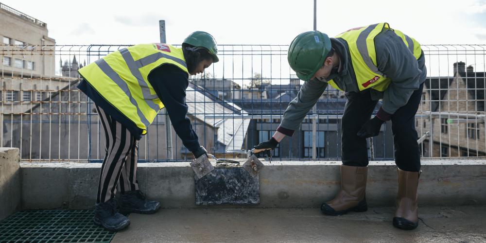 The Trinity JCR and MCR presidents put the last stone in place at the top of the Levine Building. they are both leaning over wearing hi-vis jackets and hard hats.
