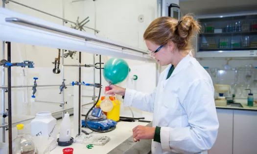 A female student in a lab coat and goggles mixes chemicals at a lab stand.
