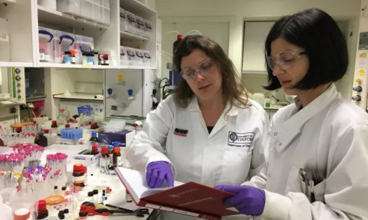 A female student in a chemistry lab consults a textbook with a researcher. Both are wearing lab coats and gloves.
