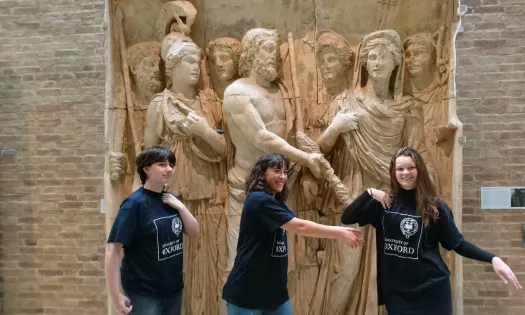 Three students wearing Oxford University T-shirts pose in front of a large sculptural wall. 