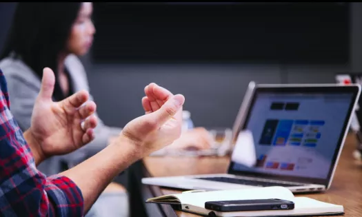 An open computer with a student's hands gesturing in the foreground; in the background is another student's head.