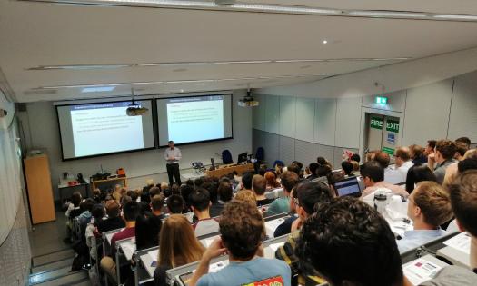 An engineering lecture viewed from the back of a small lecture theatre; at the front a lecturer is using a projection screen.