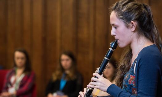 A female student plays a clarinet; in the background other students are listening.