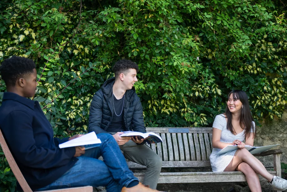 Three Trinity students sit on or next to a bench with books talking and smiling.