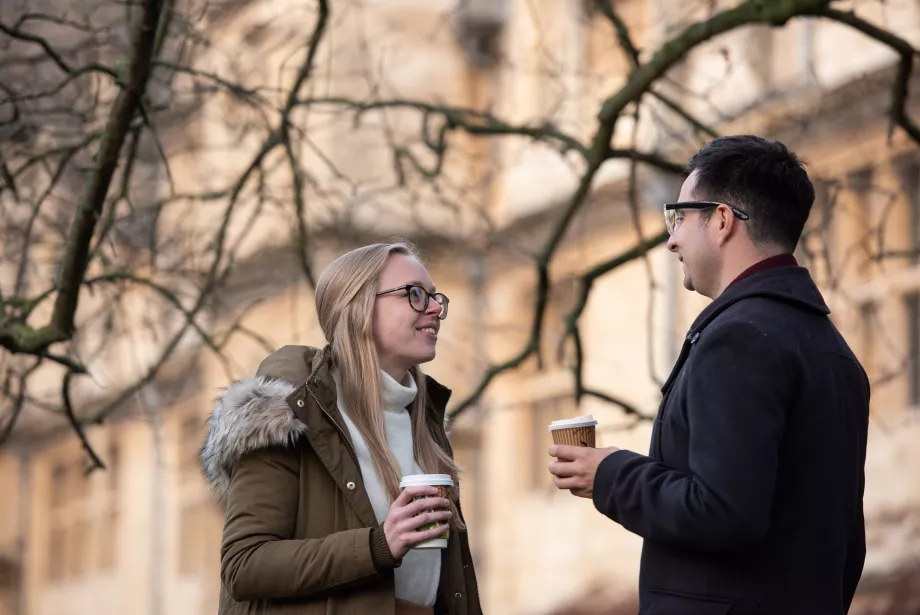 A pair of Trinity College postgraduates stand in profile holding coffee cups outside in the front quad.