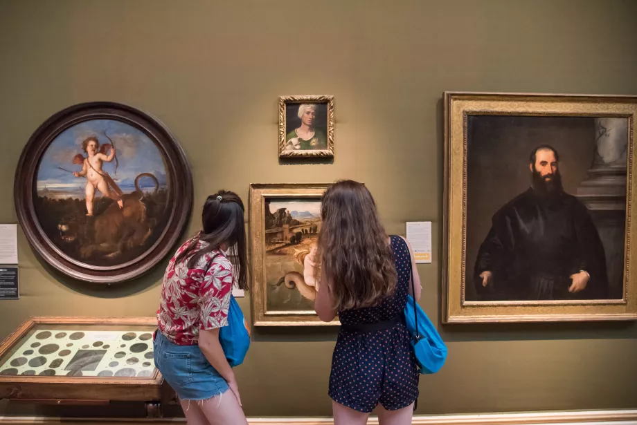 Two female students stand in a museum looking at paintings with their backs to the camera.