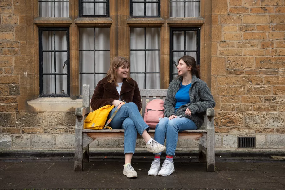 Two female students sit facing each other on a bench in Trinity and smiling as they talk.
