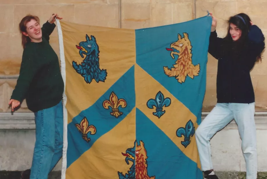 Two female students stand holding a large Trinity College flag and posing outside one of the college buildings in a photograph from the 1990s.
