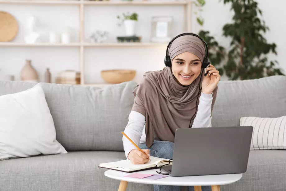 A female teacher wearing a head scarf and headset takes notes while looking at a computer during an online CPD session.