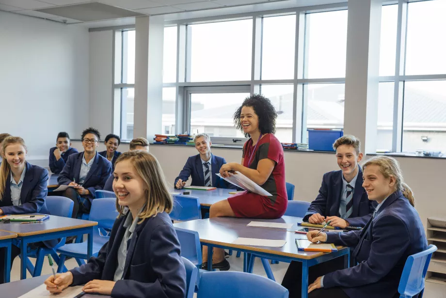 a group of sixth-form students sit at their desks in a classroom smiling at something; a teaching assistant also sits with them smiling.