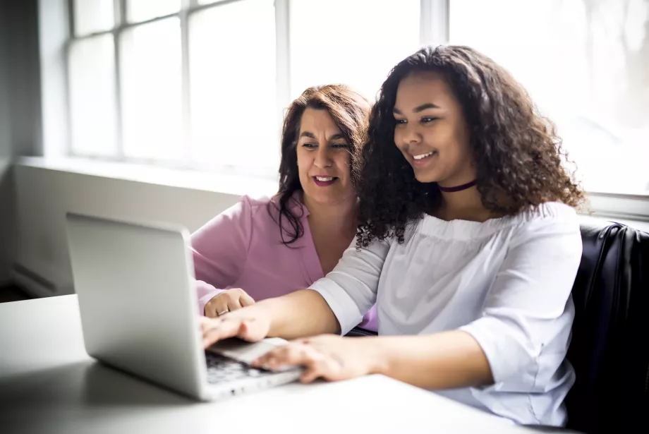 A teenage girl sits typing at a computer; her mother is crouched next to her.
