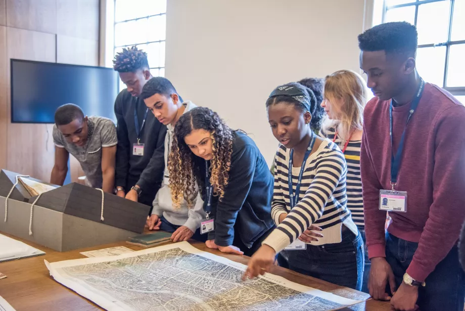 A group of students on an outreach programme wear lanyards; they are watching someone gesturing to text on a manuscript.