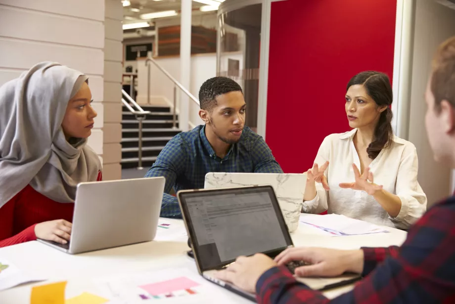 A group of students sit at a table with laptops having a discussion; one of them is wearing a head scarf.
