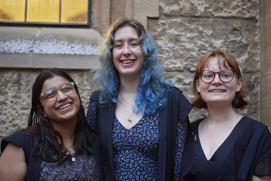 Three female students stand in gowns outside the Trinity dining hall; they are smiling.