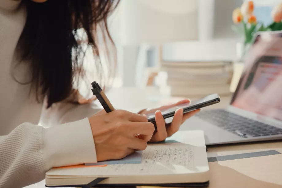 A female student sits at a computer taking notes and consulting her phone as she manages her budget.