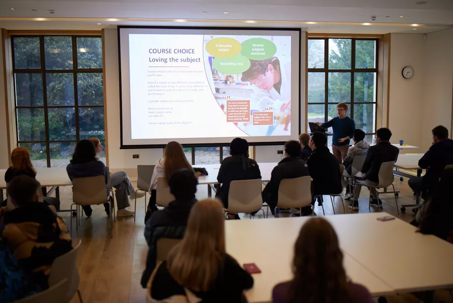 Outreach officer Rob Harrison gives a presentation talk with information on a screen to a group of secondary school students during an access visit to Trinity College.