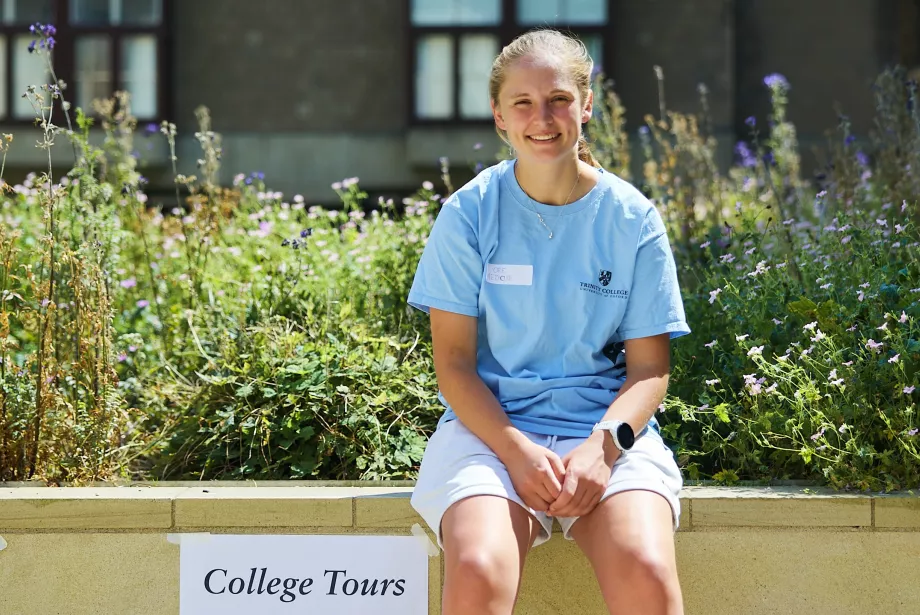 A student ambassador in a light blue t-shirt sits in Trinity's library quad next to a sign that says 'college tours leave here every 30 minutes'