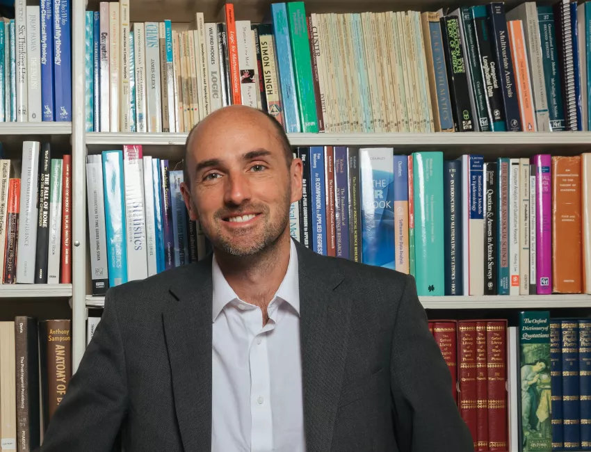 Trinity Fellow Stephen Fisher leans against a tall bookcase full of politics and sociology  books.